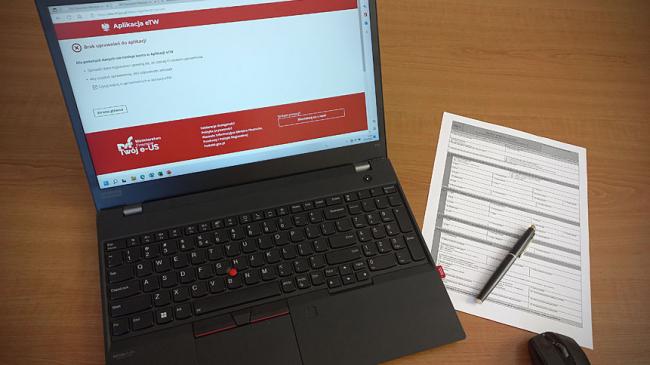 Laptop z otwartą stroną systemu Elektronicznego tytułu wykonawczego eTW (na ekranie informacja o braku dostępu), obok papierowy formularz i długopis