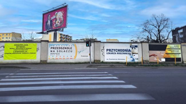 Bannery na ogrodzeniu jednej z ulic w Poznaniu, za ogrodzeniem billboard reklamowy.