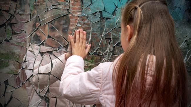 A girl touching a broken mirror with her left hand, while looking into the mirror at the same time