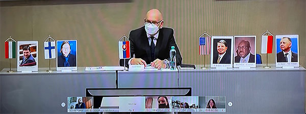 Przezes NOK Słowacji za stołem prezydialnym, obok zdjęcia i flagi reprezentujące gości łączących się on-line